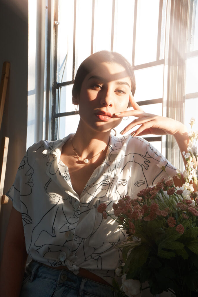 Nadya Inzunza poses by a window bathed in soft sunlight wearing a blouse with abstract faces and holding a bouquet of flowers.